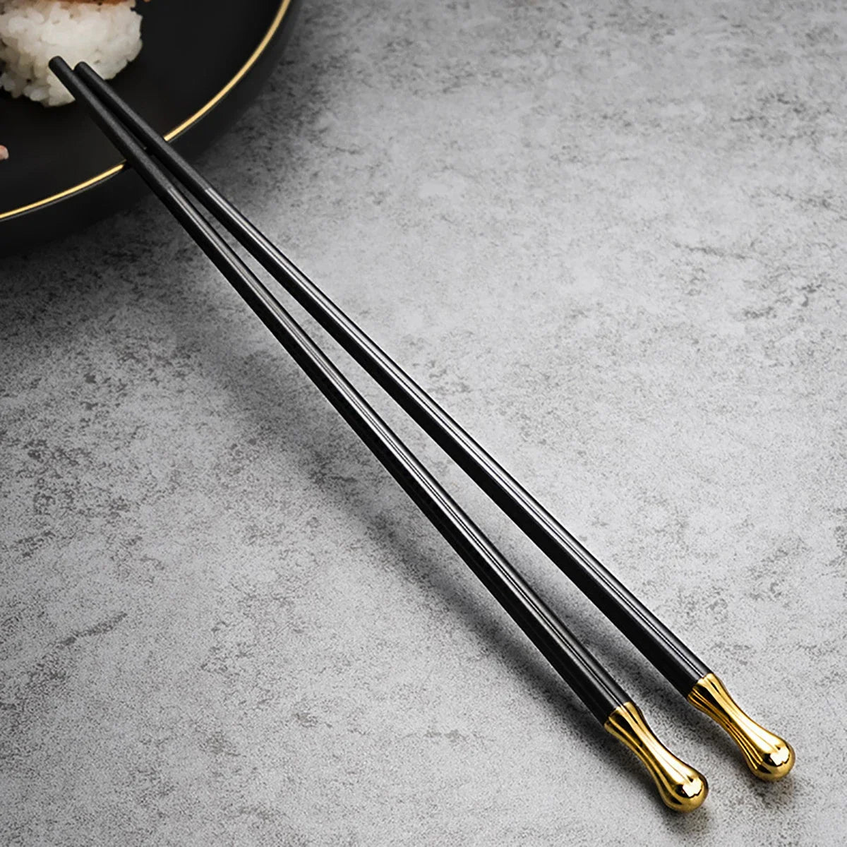 Paire de baguettes en métal noir avec embouts dorés, posée sur une table grise à côté d’une assiette noire et or contenant du riz