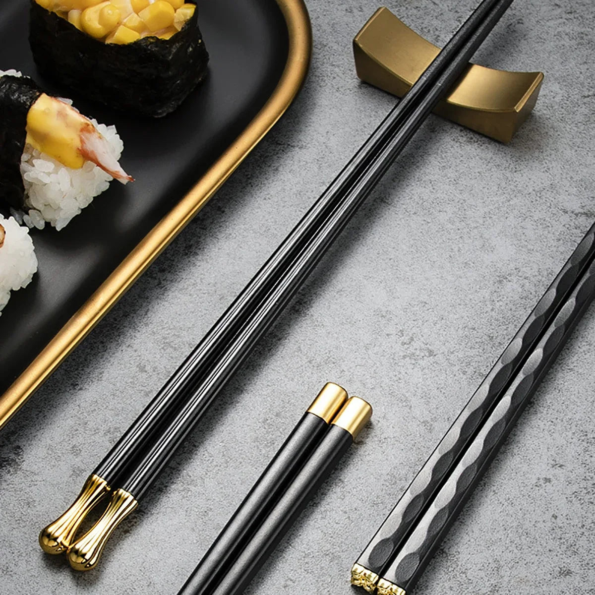 Baguettes en métal noir et doré posées avec support sur une table grise, accompagnées de sushis servis dans une assiette élégante