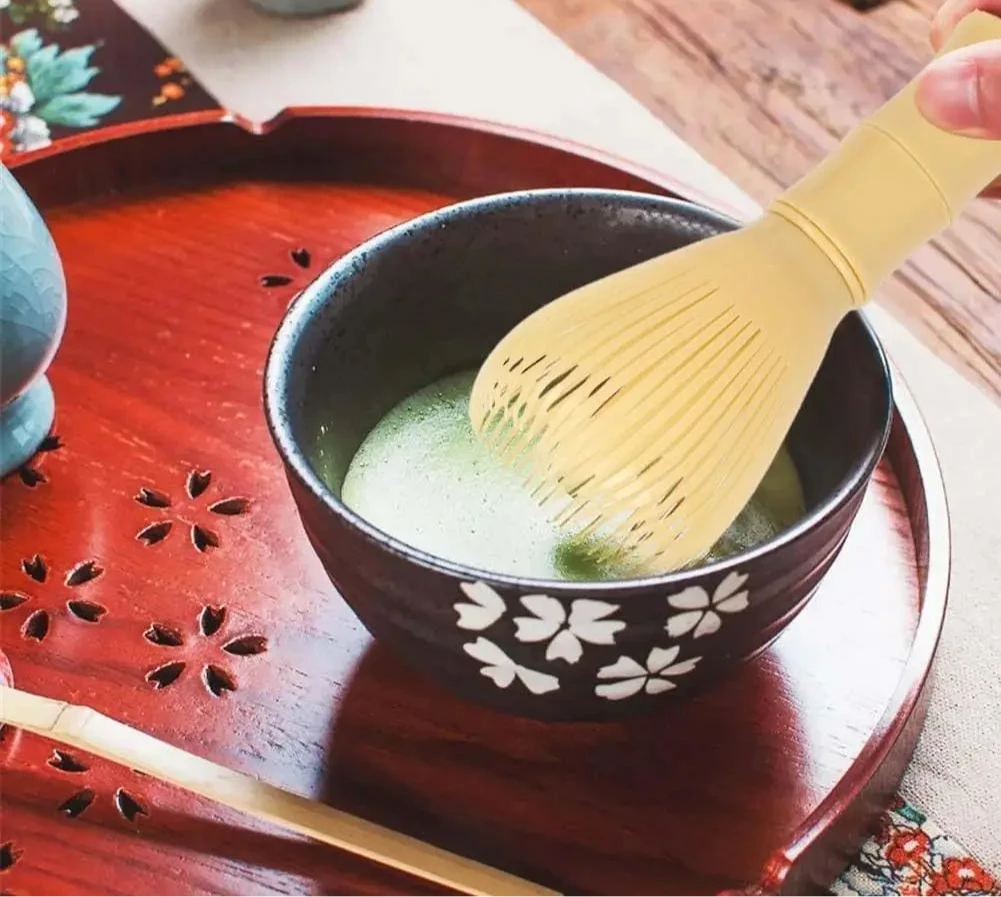 Fouet à matcha moderne jaune en train de mélanger le thé dans un bol noir fleuri, avec mousse onctueuse, posé sur un plateau rouge sculpté