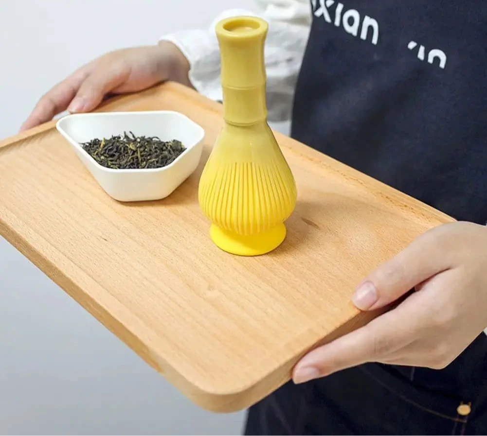 Fouet à matcha moderne jaune posé sur un plateau en bois, à côté d’un récipient de feuilles de thé, tenu par une personne en tablier