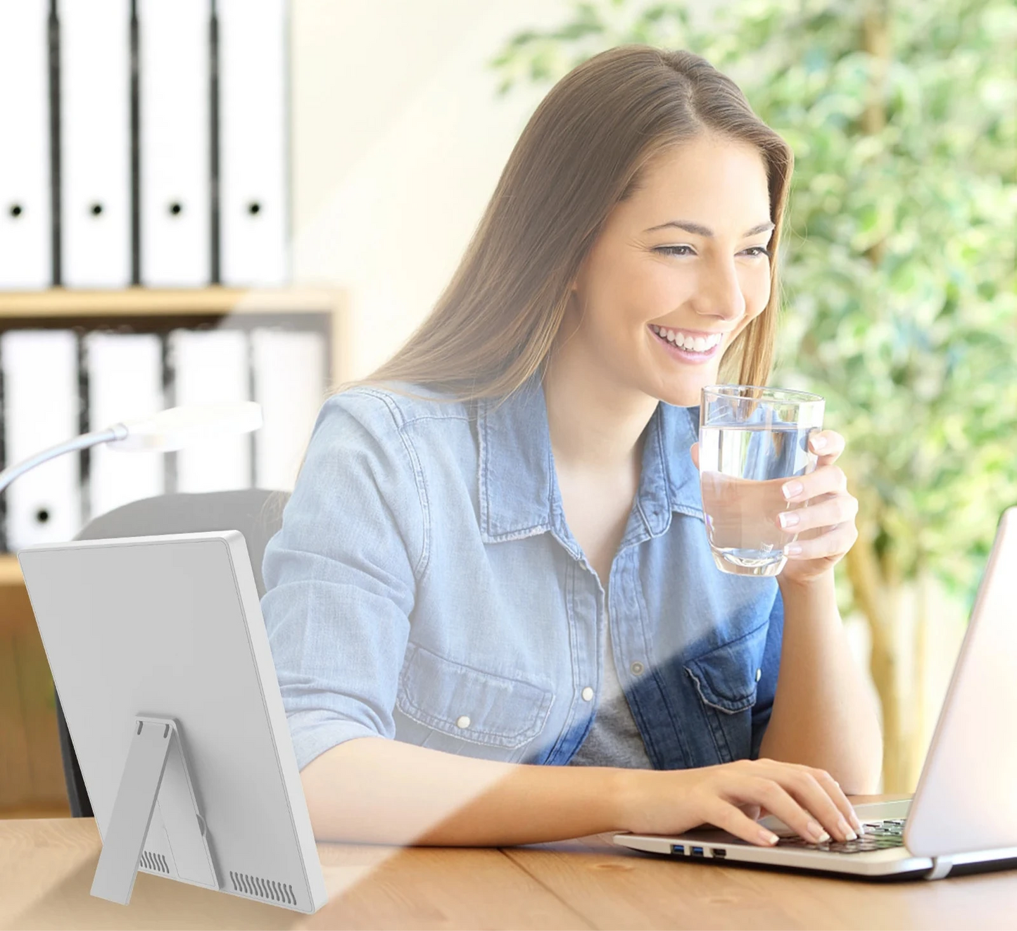 Femme souriante travaillant sur ordinateur avec une lampe de luminothérapie SAD allumée à côté – boost de lumière naturelle pour concentration et humeur au bureau.