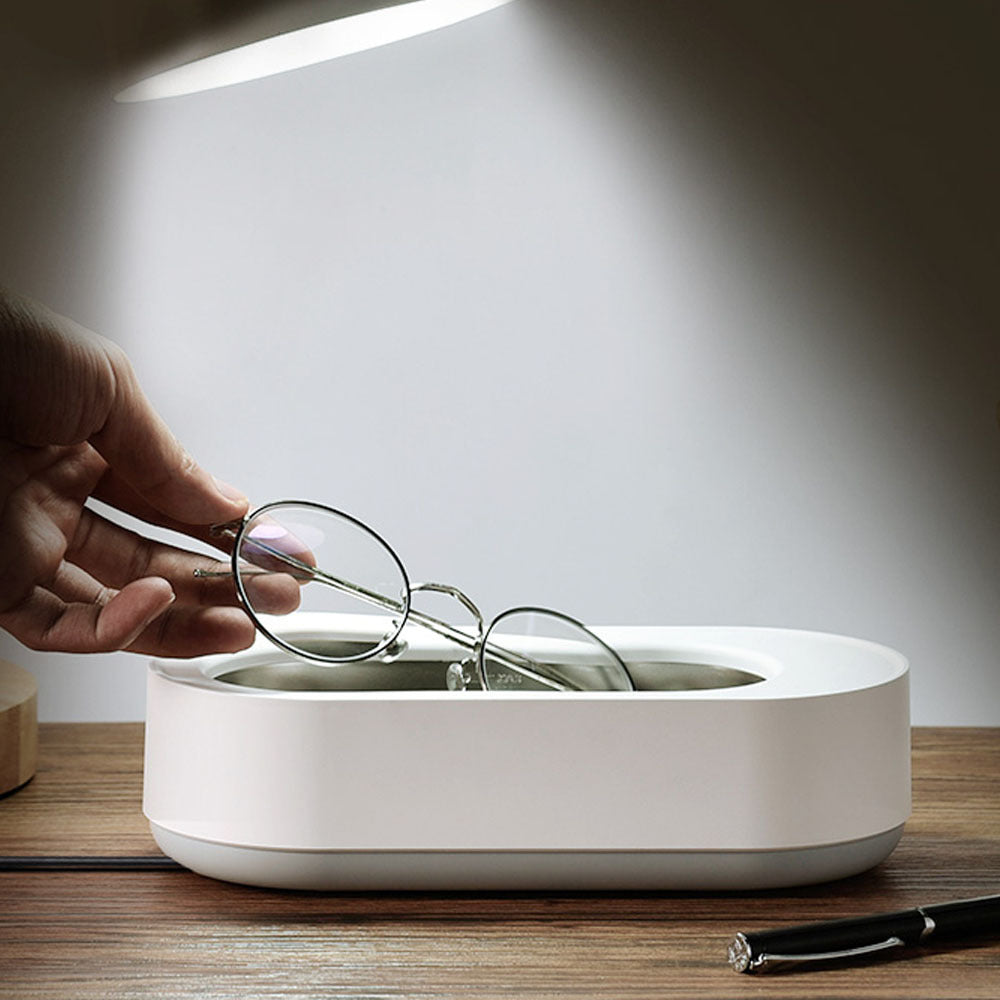 Main plaçant une paire de lunettes dans un nettoyeur ultrasonique portable 45000Hz blanc, posé sur une surface en bois, illustrant le nettoyage en douceur des verres grâce à la technologie par ultrasons