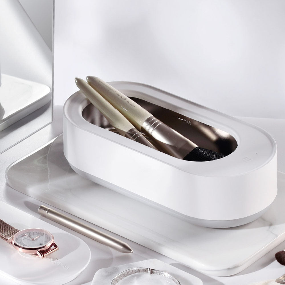Nettoyeur ultrasonique portable 45000Hz blanc en cours d’utilisation, avec plusieurs pinceaux de maquillage dans le réservoir, posé sur une coiffeuse minimaliste avec montre, stylo et bijoux à proximité.