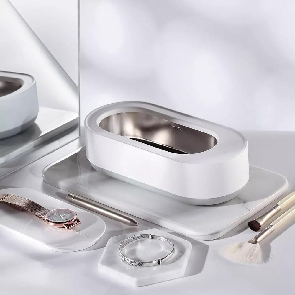 Nettoyeur ultrasonique portable 45000Hz en blanc, posé sur une coiffeuse élégante, entouré de bijoux, montre, pinceaux de maquillage et stylo, illustrant son utilisation pour un nettoyage précis et hygiénique des objets du quotidien.