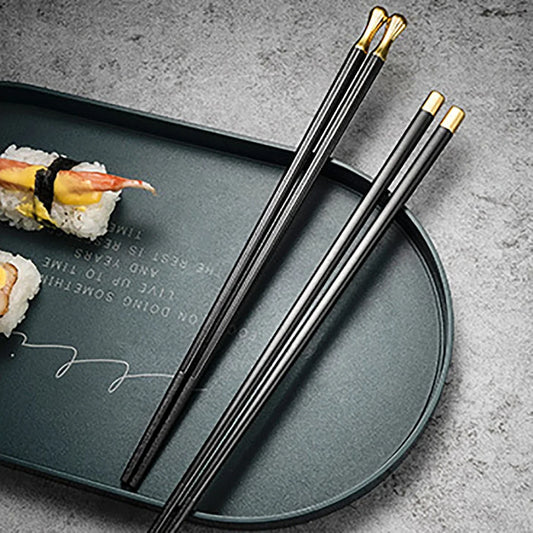 Paires de baguettes en alliage métallique noir et or posées sur une assiette à sushi, avec makis japonais en arrière-plan