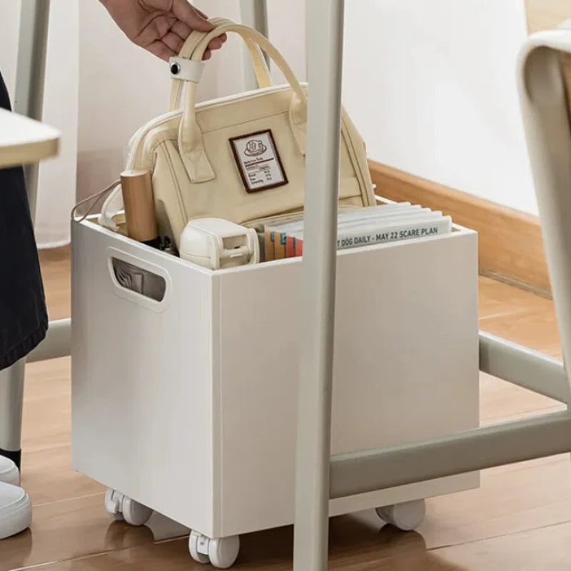 Boîte de rangement blanche à roulettes glissée sous un bureau, contenant un sac, des livres et accessoires – solution pratique et mobile pour l’organisation quotidienne.