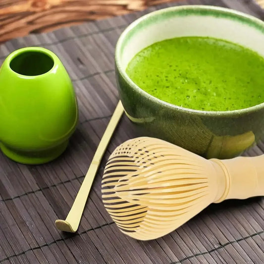 Fouet à matcha moderne posé sur une table en bambou, accompagné d’un bol de thé matcha mousseux, d’une cuillère traditionnelle et d’un pot vert assorti