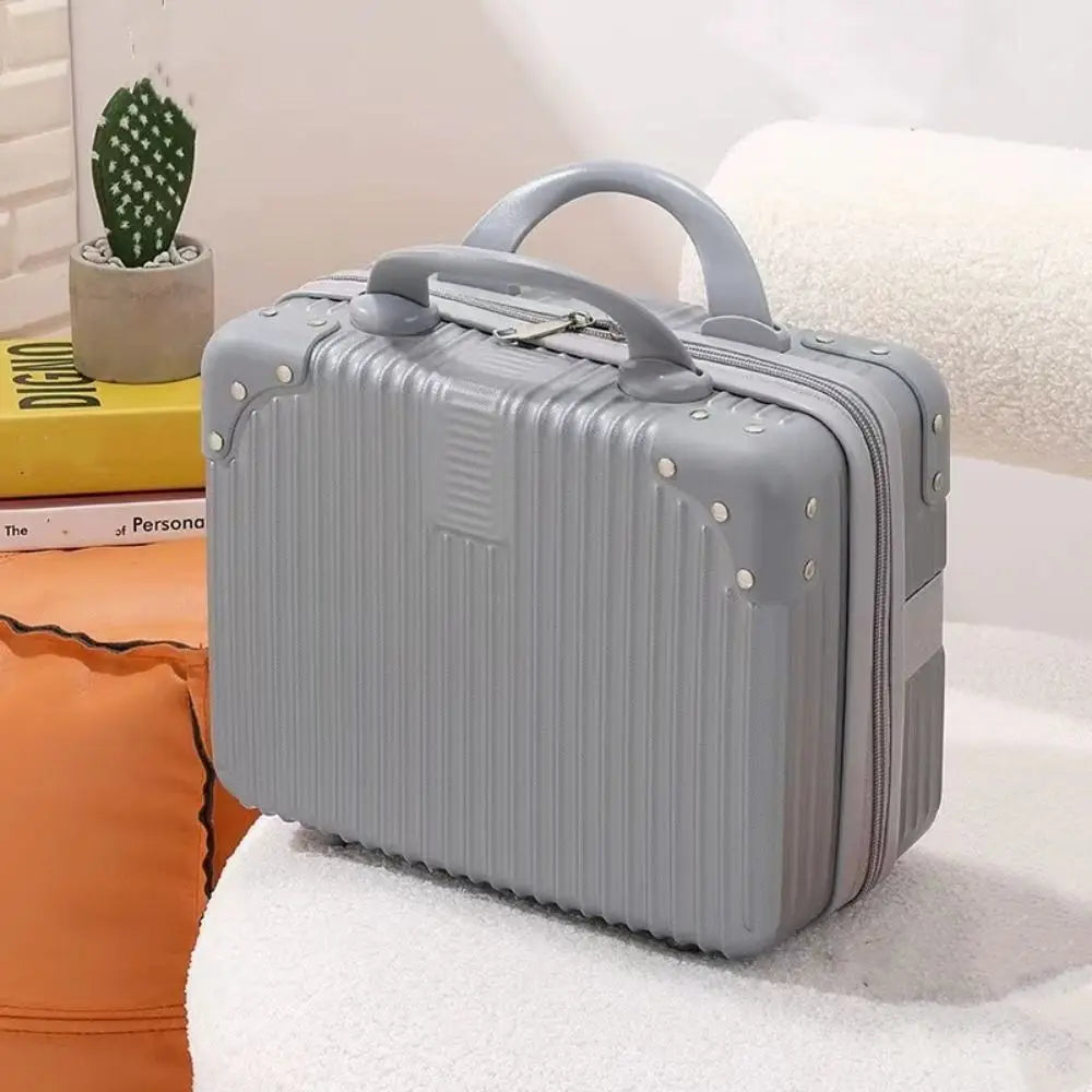 Mini valise rigide grise posée sur un canapé blanc, avec décor cosy et livre en arrière-plan, parfaite pour accessoires ou maquillage