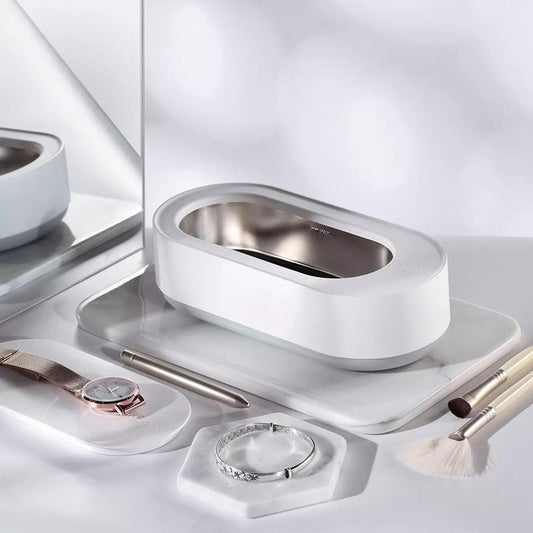 Nettoyeur ultrasonique portable 45000Hz en blanc, posé sur une coiffeuse élégante, entouré de bijoux, montre, pinceaux de maquillage et stylo, illustrant son utilisation pour un nettoyage précis et hygiénique des objets du quotidien.