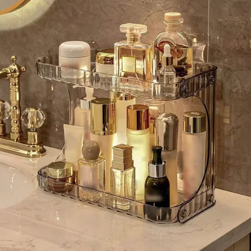 Support de rangement en acrylique transparent à deux niveaux, exposant une sélection de cosmétiques, soins et parfums dans une salle de bain élégante au style doré.